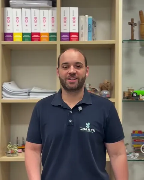 Integrante da equipe da Carletti, de camisa polo azul-marinho com o logo da empresa, em frente a uma estante com fichários de legislação tributária